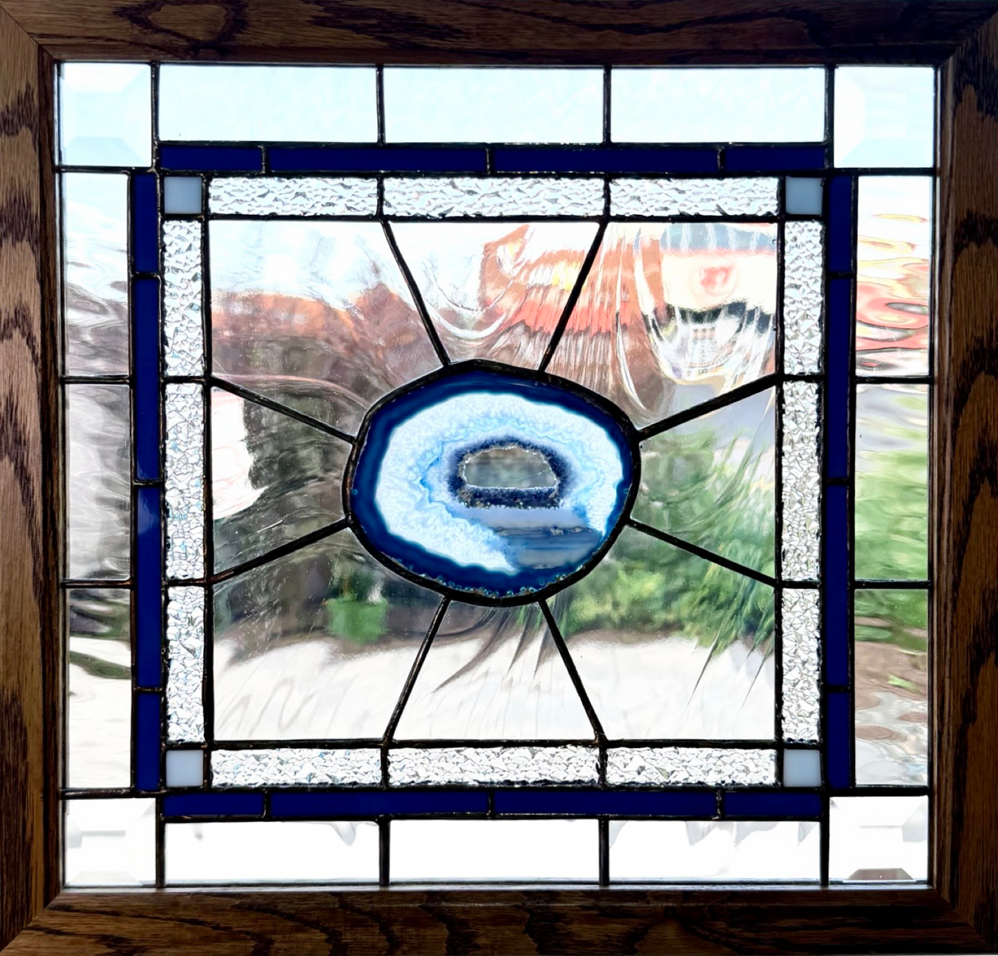 A square stained glass window panel featuring a central blue agate slice motif surrounded by textured and smooth glass sections, framed in dark brown wood with black lead lines.